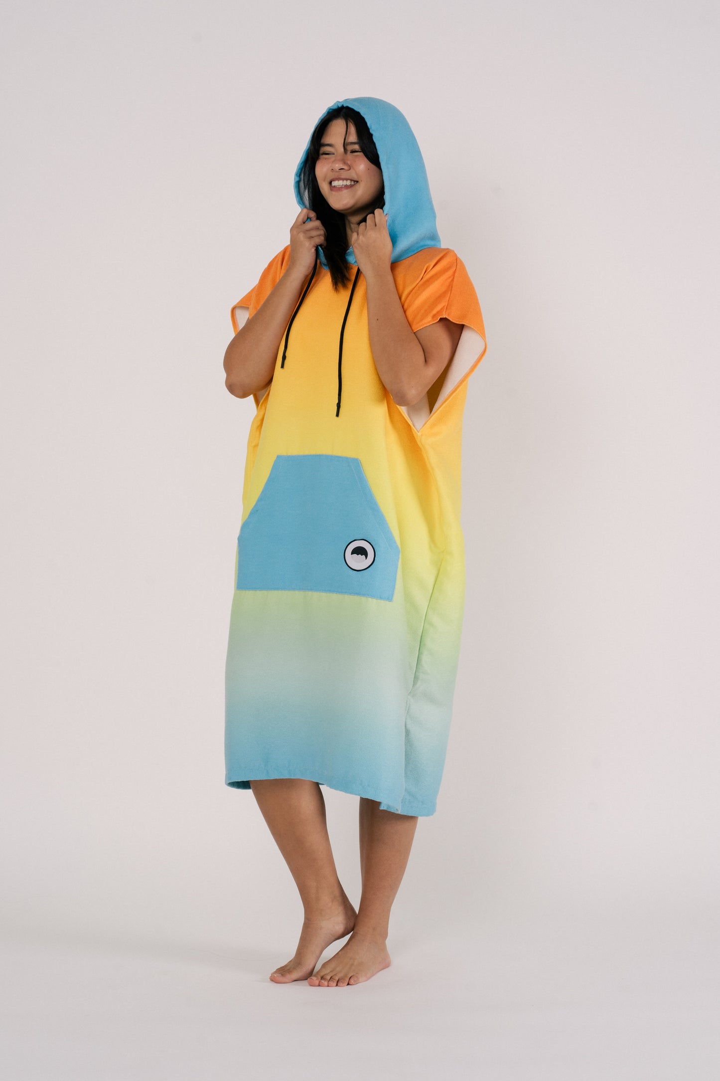 Female wearing Outwhere Changing Poncho Towel in Liwayway (dawn) design while holding hood, gradient shades of orange, turquoise, yellow, green