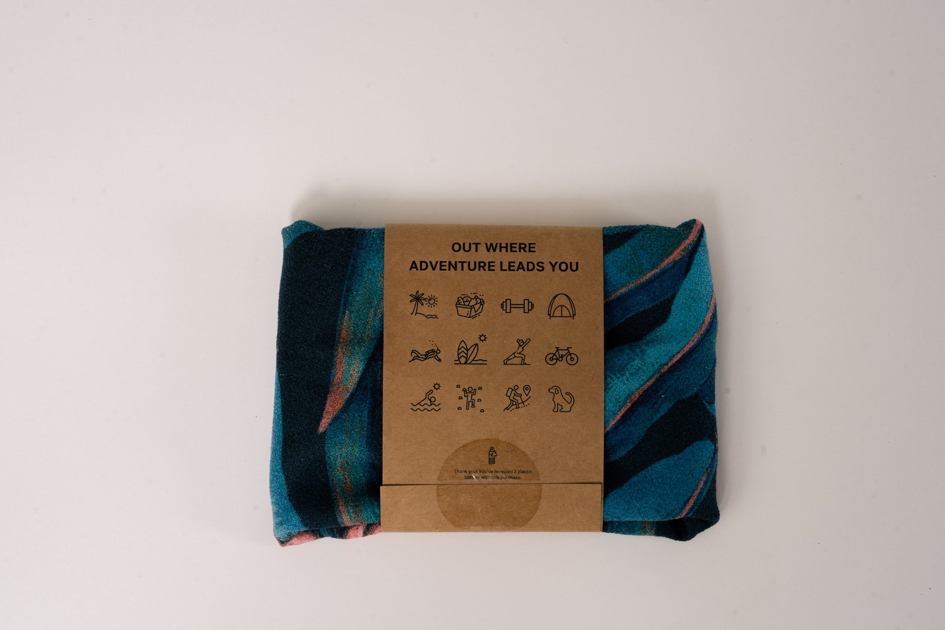 Folded Outwhere PH Everywhere Towel in Saging (banana) Blue in Packaging sleeve. Back of packaging sleeve shows tag line OUT WHERE ADVENTURE LEADS YOU, and icons that show different activities to use the towel with.